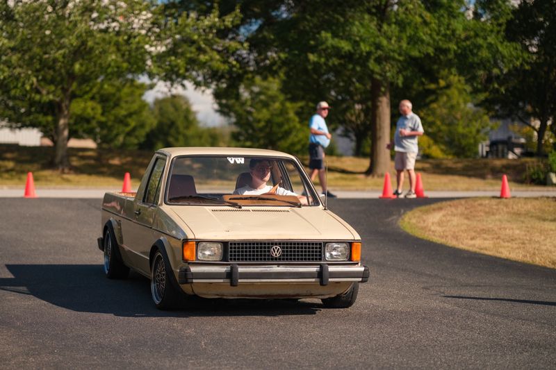 A tan 1981 Volkswagen Rabbit pickup on deep-dish wheels driving through an autocross course lined with orange cones
