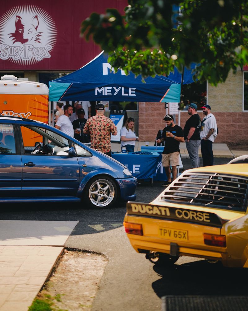 A lowered blue Volkswagen Sharan parked under a Meyle vendor tent outside the Sly Fox brewery, with a yellow Volkswagen Derby wearing a Ducati Corse livery in the foreground
