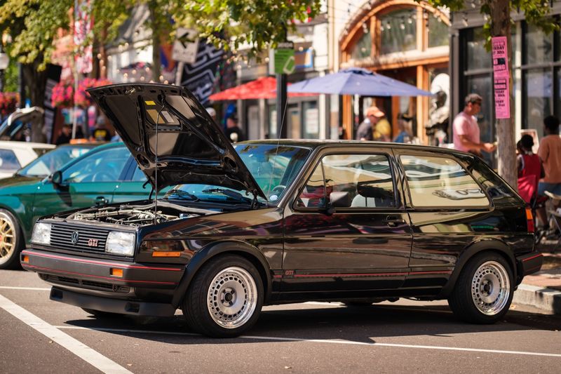 A black Volkswagen GTI Mk2 with its hood open, parked on a city street lined with shops and umbrellas