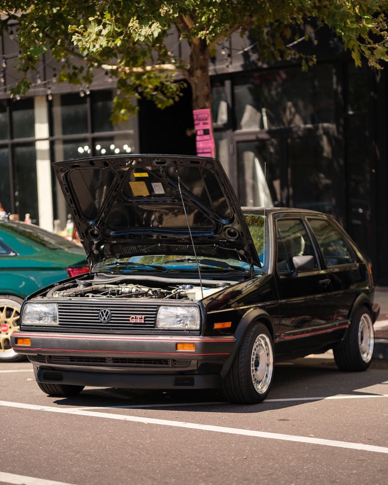 A black Volkswagen GTI Mk2 with its hood open, viewed from the front under a tree-lined street
