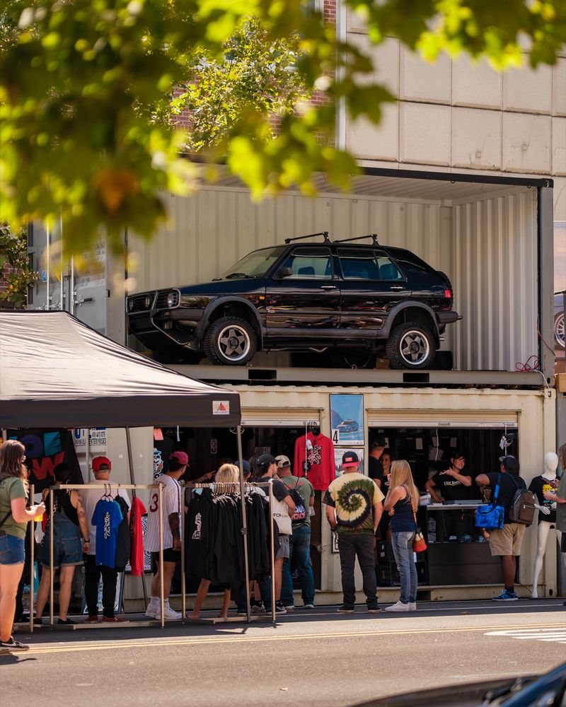 A dark green Volkswagen Golf Mk2 Syncro lifted on top of a shipping container display, with vendor stalls and shoppers below